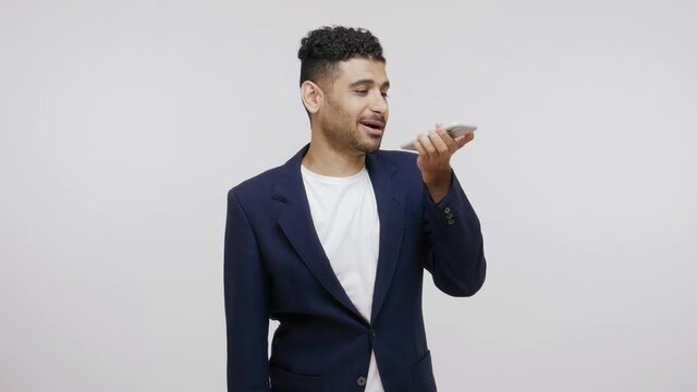 Bearded business man in suit recording voice reminder or leaving voice message holding smartphone near mouth, searching with voice using assistant app. Indoor studio shot isolated on gray background