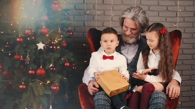 Happy family at home on Christmas Eve. Cute kids sitting on sofa with grandpa and reading book with xmas tree near. Happy holidays concept.
