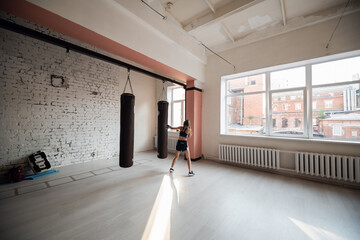 The girl is preparing for a boxing competition and trains punches on a punching bag in a spacious gym