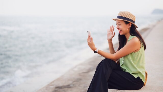 Asian traveler woman using smartphone video call online at the beach by the sea background.Concept of happy travel on vacation weekend.