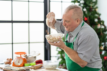 Happy senior man in kitchen apron decorating Christmas gingerbread cookies on Christmas. Chef work in the cuisine. Cook in uniform. Merry Christmas and Happy New Year. Cooking and Holiday concept