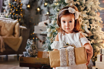 A little cute girl with a Christmas gift box near the Christmas tree, reaches out.