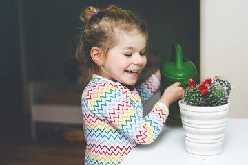 Little toddler girl watering flowers and cactus plants on window at home. Cute child helping,...