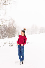 Fototapeta na wymiar A young beautiful girl in a red sweater stands among the snowdrifts in the forest. Woman in black cap in a park winter when it snows.