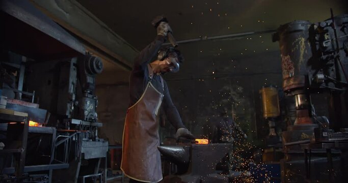 In general, in an atmospheric forge, a blacksmith powerfully strikes the red-hot metal with a huge hammer, sparks fly off. The man's face is black with soot and wears a leather apron.