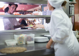 Smiling waitress taking cooked ordered meals in restaurant kitchen for serving guests