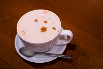 close-up of a white cup of coffee with froth decorated with cinnamon stars on a saucer with a spoon standing on a wooden table
