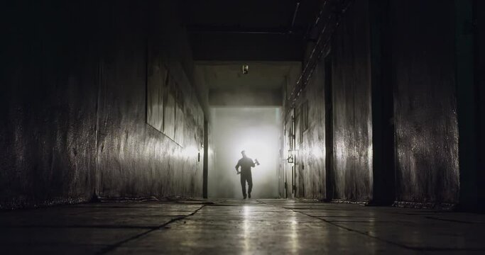 A silhouette of a man with a large ax emerges from the white fog, confidently walking towards the camera along a dark, gloomy and scary corridor. High quality 4k footage.