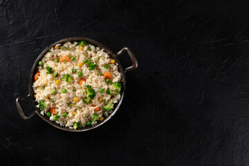 Vegan rice in a pan, top-down shot on a dark background with copy space. Healthy dinner, cooked with broccoli, green peas and carrots