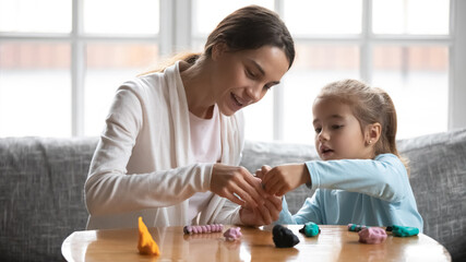 Obraz na płótnie Canvas Caring young mother making plasticine figures with little baby daughter, developing creativity at home. Affectionate nanny helping small girl, involved in handmade activity indoors, daycare concept.
