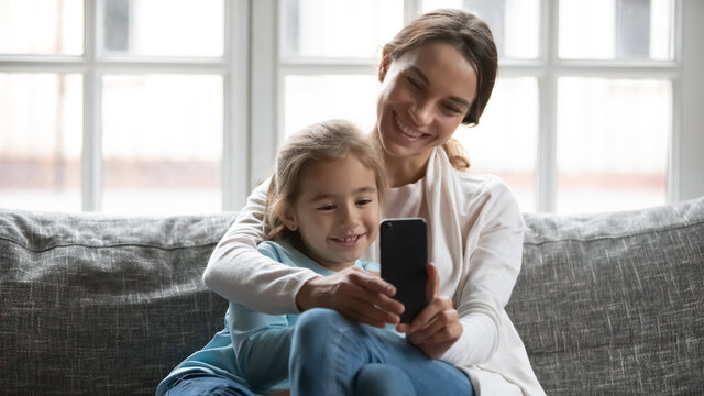 Smiling beautiful mother cuddling adorable pretty little kid daughter, using smartphone together at home, posing for selfie photo or recording funny video for social networks, shopping online.
