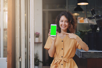 Mockup image of a woman wearing yellow dress holding and showing mobile phone with blank green screen at coffee shop