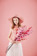 Portrait of smiling young pretty woman in straw hat holding branches of blooming oriental cherry
