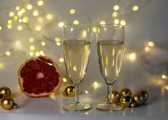 two glasses of champagne, pink grapefruit, golden glass balls and various Christmas decorations, illuminated background of a chain of white lights, Christmas waiting time