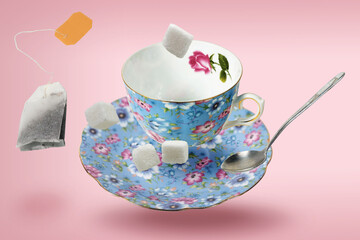 Decorated empty blue porcelain teacup with saucer, tea bag, spoon and sugar cubes flying over pink surface