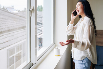 Fototapeta na wymiar Happy young Asian woman laughing and talking on phone with friend when standing at apartment window