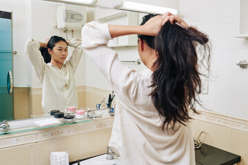 Pretty young Chienese woman in white shirt making high ponytail in front of mirror in bathroom