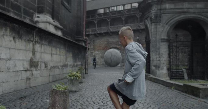 Side view of teenager soccer player kid kicking football ball with knee. Talanted boy practicing tricks at old city street. Concept of sport and lifestyle. Person on bike at background