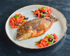 Fried fish with fresh herbs, tomatoes and olives served on a white plate over black rustic wooden table background.