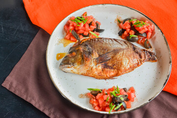 Fried fish with fresh herbs, tomatoes and olives served on a white plate over black rustic wooden table background.