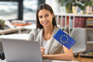 Beautiful woman student with EU flag sitting at table with laptop and smiling while looking at camera. EU education concept