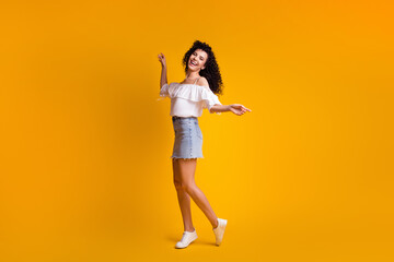 Full size photo of pretty cheerful brunette woman dancing wear white top blue skirt sneakers isolated on yellow color background