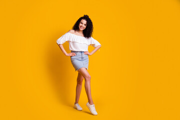 Fototapeta na wymiar Full size photo of cheerful flirty brunette woman standing hands on hips wear white top blue skirt sneakers isolated on yellow background