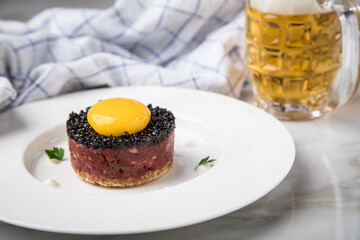 Gourmet Rindertatar Beef Tatar roh aus Filet fettarm, Kapern, Eigelb, Zwiebeln auf Toast Brot und schwarzer Kaviar mit Bier, Küchentuch und Marmor Hintergrund hell