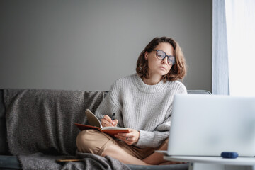 Beautiful young girl student studying at home online while sitting in a warm sweater on the couch...