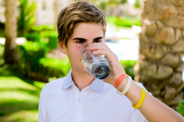 Young caucasian boy drinking clear fresh mineral water, outdoors during summer time. Health concept.