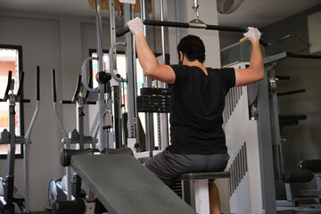Sporty man wearing protective face mask exercising with lat pulldown machine to build arm and shoulder muscle in fitness gym. Bodybuilding new normal concept.