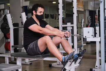 Fitness young man wearing protective face mask using rowing machine in the gym. New normal exercise.