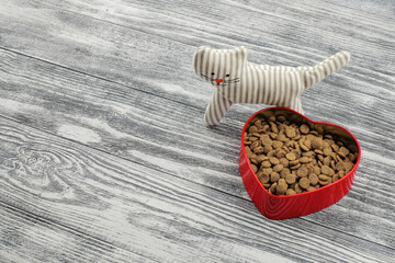 Food for pets in a red heart shaped bowl and toy on wooden background. With love for cats. Copy space.