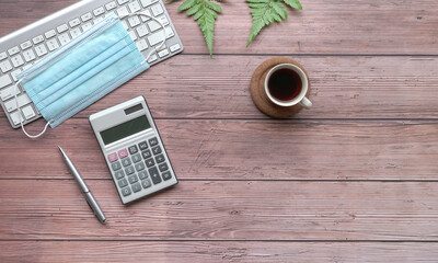Wood table with keyboard, face mask, coffee, calculator and green plants.