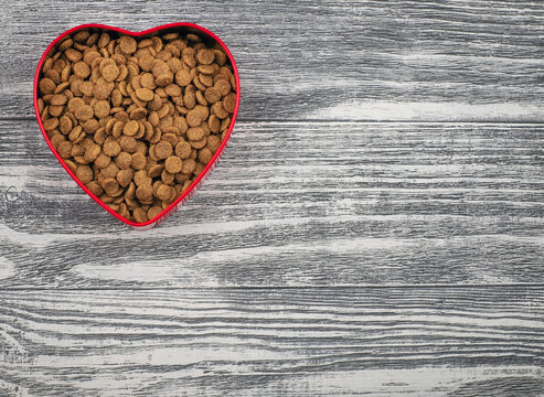 Red heart shaped bowl with food for pets on wooden background. With love for animals concept and Valentine's Day. Copy space.