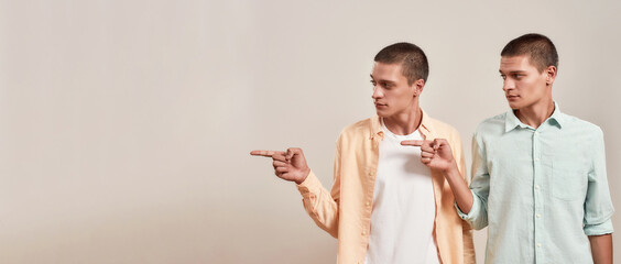 Two young twin brothers in casual wear pointing to the left and looking aside while standing together isolated over beige background - obrazy, fototapety, plakaty