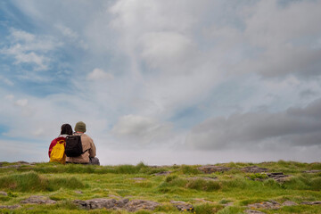 Man and woman tourists with backpacks sitting close to each other on a green grass, looking at a beautiful blue cloudy sky, their back to camera. Concept love and mutual hobby and interests.