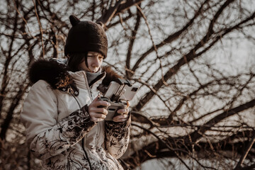A girl with a phone is standing in a hat and jacket against the background of trees.