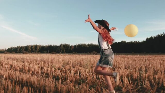 Golden-haired girl in denim overalls and a cowboy hat with a yellow balloon in her hand runs through an agricultural field. Happy child is playing among the straw rolls. Concept of youth and freedom