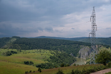 Obraz premium High voltage transmission lines and towers runing trough green lawn and forest on mountain Uvac, Zlatar with blue sky and meanders in the background.Meanders of river Uvac, mountain Zlatar, Serbia