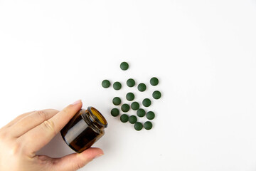 A female hand holds a jar of pills, near scattered green spirulina pills on a white background.