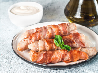 Ready-to-eat pigs sausages wrapped in bacon on plate. Fried savory sausages wrapped in bacon served fresh green basil leaves with sauce on background. Shallow DOF. Copy space for text.