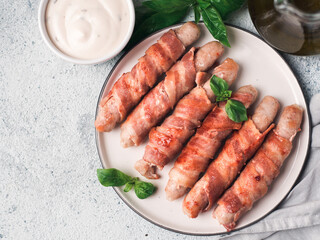 Ready-to-eat pigs sausages wrapped in bacon on plate. Fried savory sausages wrapped in bacon served fresh green basil leaves with sauce on background. Copy space for text. Top view or flat lay.