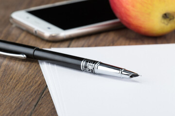fountain ink pen with cap on table with sheets of white paper and phone with red apple