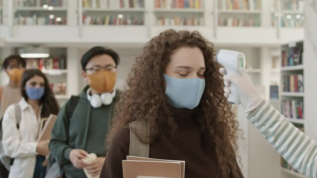 Multicultural university students wearing cloth facial masks standing in line in library, moving forward, unrecognizable hand measuring their body temperatures with contactless thermometer
