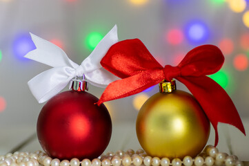 Christmas decorations: a red ball with a white bow and a gold ball with a red bow, lying in pearls on a white wooden background with blurred Christmas tree lights in the background.