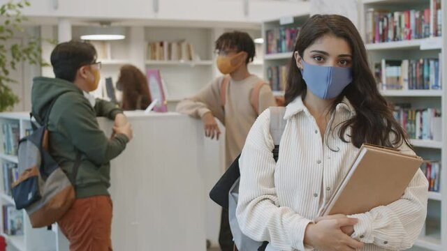Medium POV of Mixed-Race brown-eyed female student wearing custom-made face mask, holding books, standing in library, looking on camera. Diverse people talking, walking behind