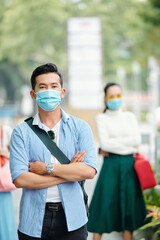Fototapeta na wymiar Handsome confident young Vietnamese man in medical mask standing outdoors with arms folded and looking at camera