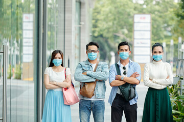 Unhappy serious young Vietnamese people in medical masks standing in street with arms folded and looking at camera