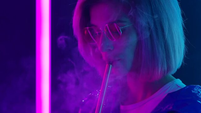 Portrait of a young stylish woman in sunglasses smoking a hookah and blowing out puffs of smoke. Close up. Slow motion.